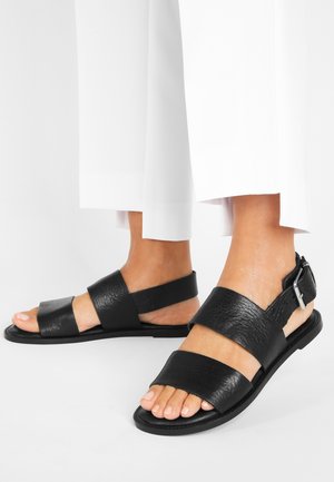 Voeten in zwarte leren sandalen met gespen en twee brede bandjes, gecombineerd met witte cropped broek, staand op een witte vloer.