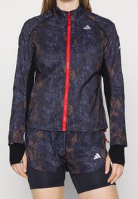 Kvinde iført en mørkeblå og brun mønstret zip-up jakke med rød lynlås og matchende shorts, lagd ind over sorte kompressionsshorts.