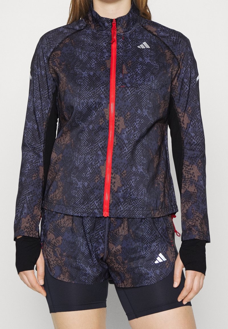 Kvinde iført en mørkeblå og brun mønstret zip-up jakke med rød lynlås og matchende shorts, lagd ind over sorte kompressionsshorts.