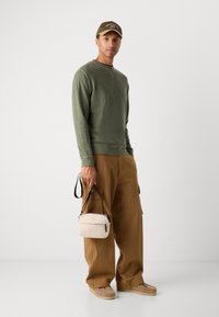 Olijfgroene sweater, kaki cargobroek, beige schoudertas met een zwarte rits en lichtbruine instapschoenen. Eenvoudig ontwerp en relaxte pasvorm.