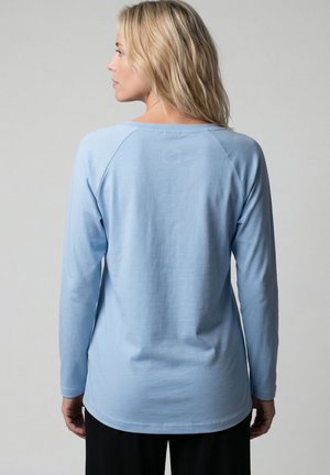 Frau in hellblauem Langarmshirt, Rückenansicht, mit blondem Haar und schwarzer Hose vor schlichtem Hintergrund.