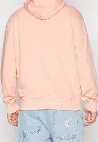 Pfirsichfarbene Kapuzenjacke mit bequemer Passform, langen Ärmeln und einer Kängurutasche. Verfügt über gerippte Bündchen und Saum sowie ein Logodetail an der Seite.
