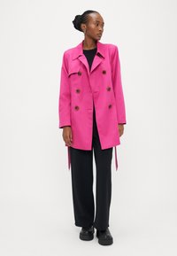 Ροζ παλτό τύπου trench με έξι καφέ κουμπιά, ωμοπάζα και πλευρικούς δεσίματα, φορεμένο πάνω από μαύρο τοπ και παντελόνες με φαρδιά πόδια, συνδυασμένο με χοντρά παπούτσια.