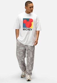 Bílé oversized tričko s barevným motivem Mickey Mouse a černým textem, v kombinaci se světle šedými tečkovanými sportovními kalhotami a teniskami.