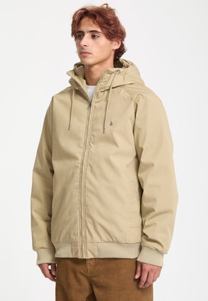 Jeune homme portant une veste à capuche beige zippée avec des poignets côtelés et un pantalon en velours côtelé marron, debout devant un fond blanc uni.