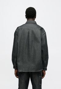 Ciemnoszara koszula dżinsowa o luźnym kroju, zapinana na guziki, z mankietami oraz kontrastującym białym przeszyciem. Widoczna zakładka z tyłu dodaje faktury i głębi.