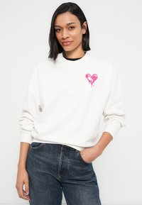 Λευκό φούτερ με γραφικό ροζ καρδιάς που στάζει στη αριστερή πλευρά του στήθους, συνδυασμένο με σκούρο μπλε τζιν. Μαλακό ύφασμα, άνετη εφαρμογή.