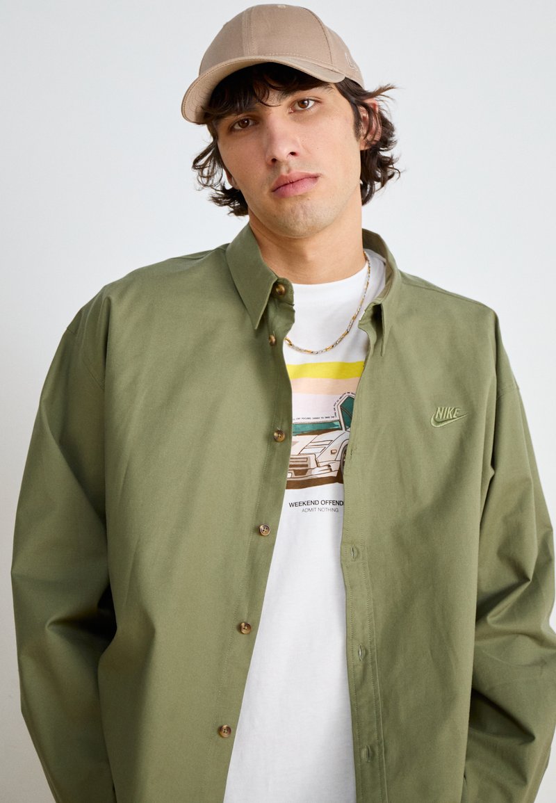 Camisa de botones verde oliva de corte relajado, combinada con una camiseta blanca con un diseño de coche y una gorra beis.