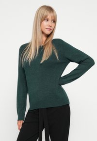 Mørkegrønn strikket genser med rund halslinning, lange ermer og ribbestrikket kant, kombinert med svarte bukser med belte. Glatt tekstur, uformelt design.