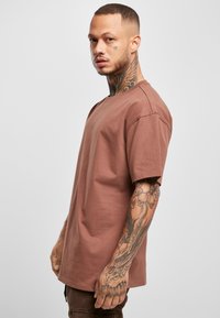 Brun bomull T-skjorte med løs passform, rund hals og korte ermer; modellen viser tatoveringer og står mot en enkel bakgrunn.