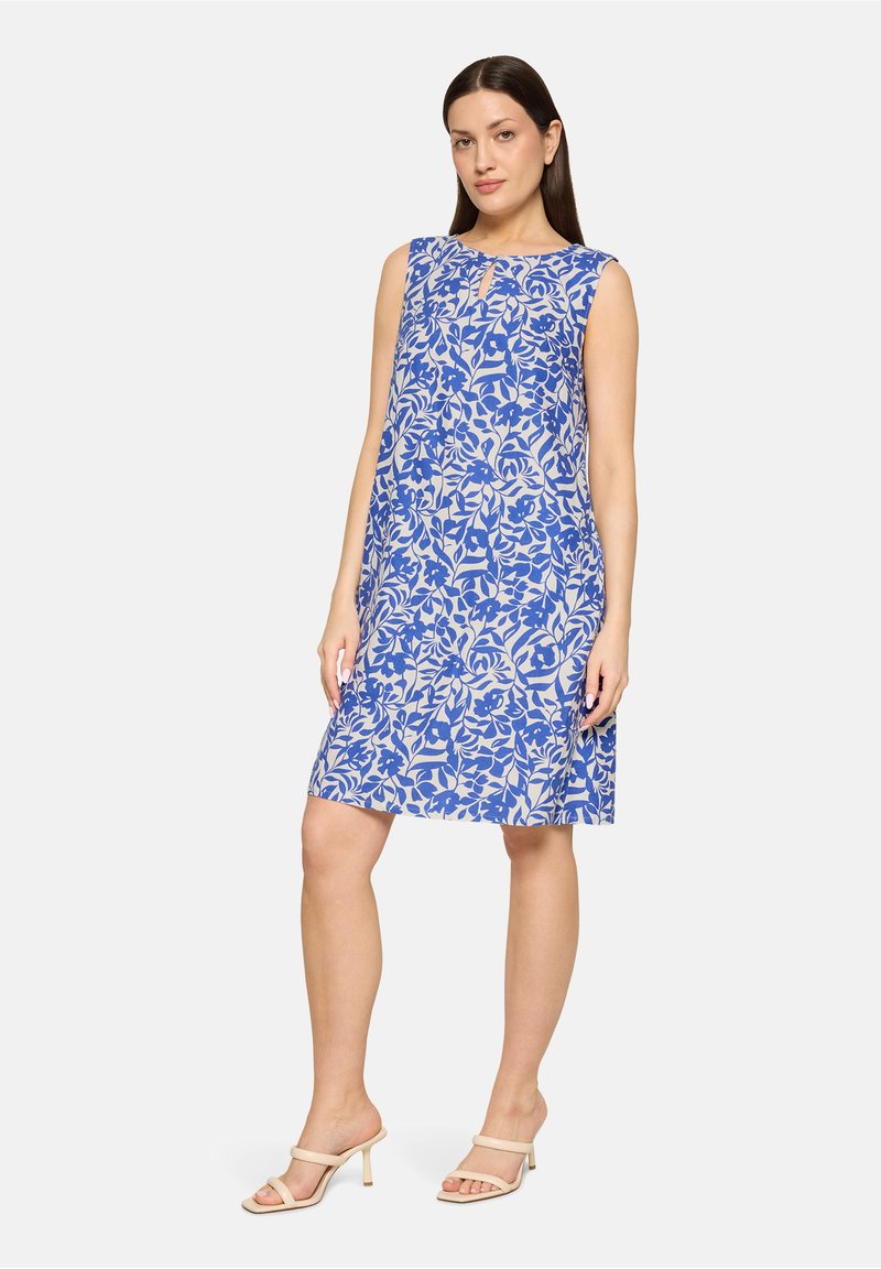 Femme debout portant une robe sans manches longueur genou avec des motifs floraux bleus et des sandales beiges à talons sur un fond uni.