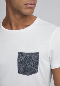 Camiseta blanca de cuello redondo con un bolsillo de parche oscuro de diseño estampado, que presenta flores y patrones de paisley. Tejido suave, forma ajustada, sin más adornos.