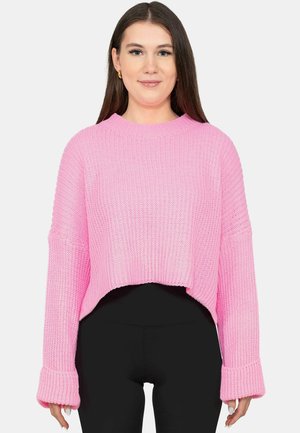 Frau trägt einen lockeren, gerippten rosa Pullover mit langen Ärmeln über schwarzen eng anliegenden Hosen, sie steht vor einem schlichten hellen Hintergrund.