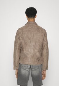 Giacca in suede marrone con colletto, dettaglio sul retro e polsini con bottone, abbinata a pantaloni corti grigi. Finitura testurizzata su tutta la giacca.