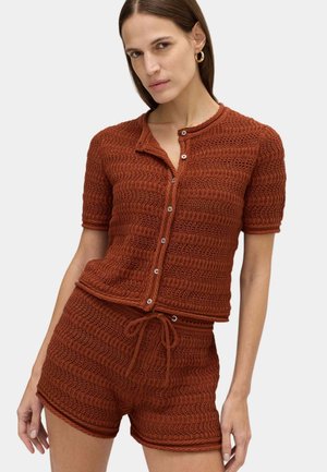 Frau, die ein rostfarbenes gestricktes Kurzarm-Button-up-Oberteil und passende Shorts mit Kordelzug trägt, stehend vor einfachem Hintergrund.