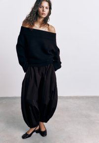 Vrouw met los krullend haar, een zwart off-shoulder trui, volumineuze zwarte broek en zwarte slingback ballerina's, staand op een betonnen vloer.