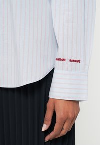 Zbliżenie na dłoń obok mankietu jasnoniebieskiej koszuli z czerwonymi pionowymi paskami i czerwonym haftowanym napisem „SAMSØE SAMSØE”, noszonej na czarnej plisowanej spódnicy.