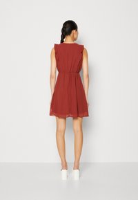 Kobieta stojąca tyłem, ubrana w bezrękawową sukienkę w rdzawym kolorze z fakturą i falbanami, na białych platformach, na jednolitym tle.