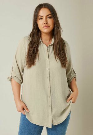 Chemise beige clair à manches longues avec une finition texturée, devant à boutons et manches retroussées. Associée à un jean bleu, avec un ourlet arrondi.
