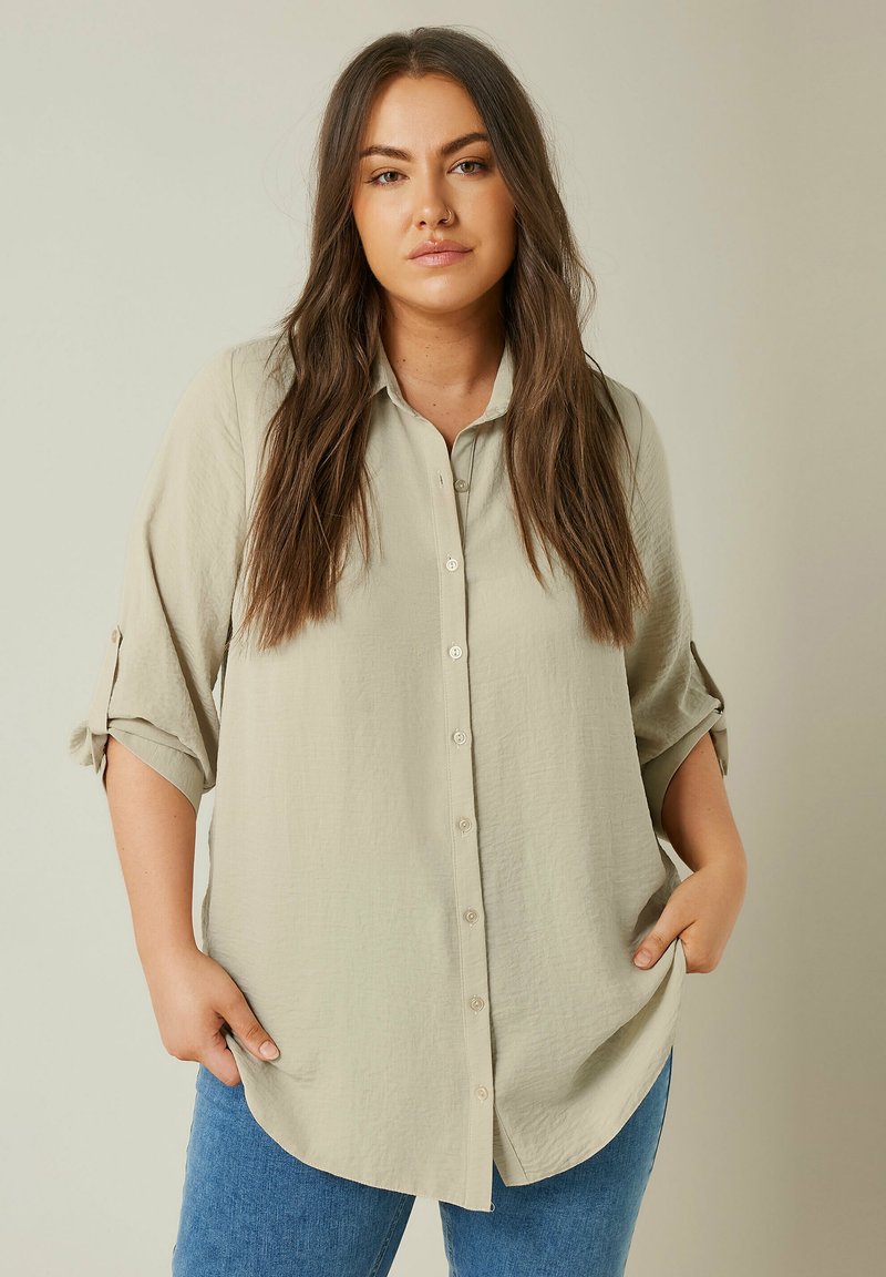Helles beiges Langarmshirt mit strukturiertem Finish, Knopfleiste vorne und hochgekrempelten Ärmeln. Kombiniert mit blauen Jeans, die einen geschwungenen Saum haben.