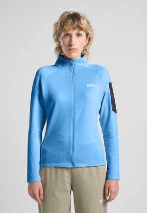 Giacca azzurra chiara con zip, texture a coste, colletto alto e tasca nera accentata sul braccio sinistro. Abbinata a pantaloni khaki.