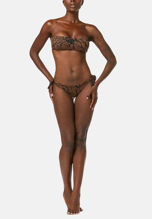 Femme debout pieds nus en bikini deux-pièces marron avec motif abstrait noir, mains posées sur les hanches sur fond blanc uni.