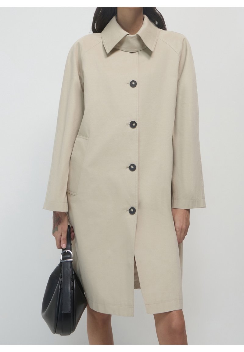 Femme portant un manteau beige boutonné jusqu'aux genoux, tenant un sac à main noir dans sa main gauche sur un fond uni.