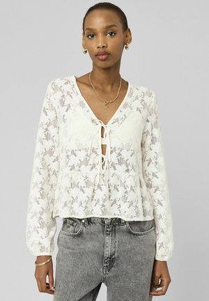 Femme portant un chemisier à manches longues en dentelle fleurie blanche avec des liens à l'avant, un jean taille haute gris et des bijoux en or, debout devant un fond gris clair.