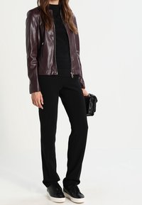 Bordeaux lederen jack met hoge kraag, zwart aangesloten top, zwarte broek en zwarte sneakers. Houdt een kleine zwarte handtas vast.