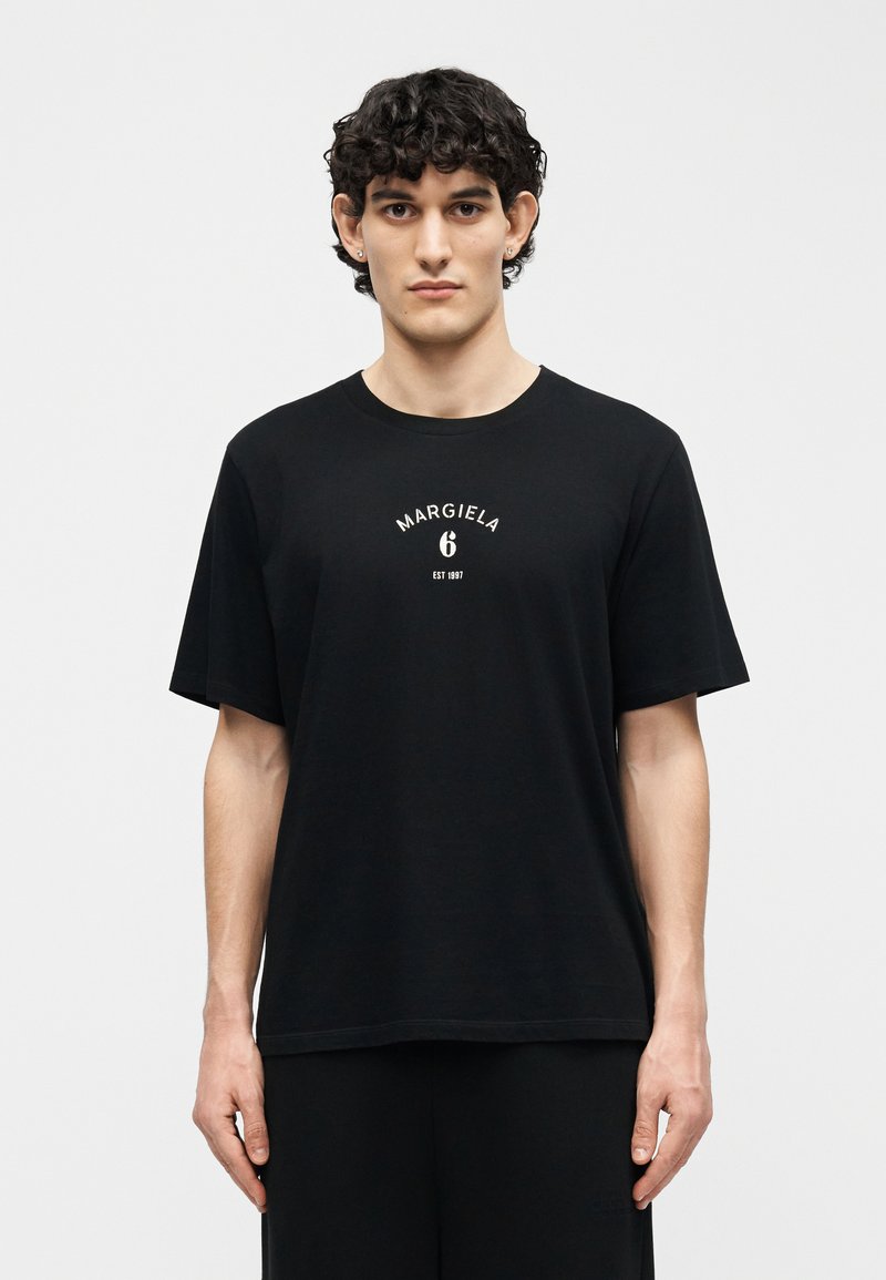 Νέος άνδρας με σγουρά σκούρα μαλλιά, φορώντας μαύρο T-shirt Margiela 6 και μαύρο παντελόνι, στέκεται μπροστά σε ένα απλό φωτεινό φόντο.