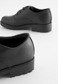 Chaussures en cuir noir avec un bout arrondi et une semelle en caoutchouc texturé, dotées de coutures discrètes et d'un talon bas.