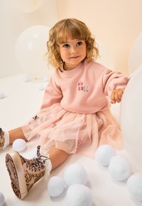 Felpa rosa con dettagli di paillettes e gonna in tulle. Tessuto morbido e strutturato. Stivaletti alla caviglia color oro con glitter e dettagli in pizzo. Pompon bianchi e blu.