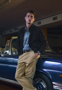 Giacca di suede marrone sopra una camicia di denim blu, pantaloni khaki e un orologio d'argento. Appoggiato a una classica auto cabriolet blu.