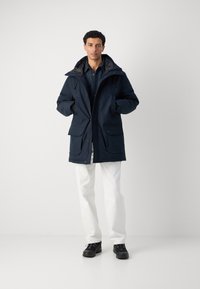 Navy blue parka with a hood, multiple front pockets, and a zip closure. Features a smooth exterior and a lined interior for warmth. White pants.
