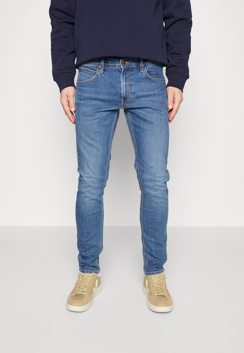 Homme portant un jean slim bleu, des baskets beige et un haut à manches longues foncé, debout contre un fond clair uni.