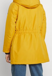 Chaqueta acolchada amarilla con capucha, cintura elástica y acentos negros, con una textura suave y un corte recto a la altura de la cadera.