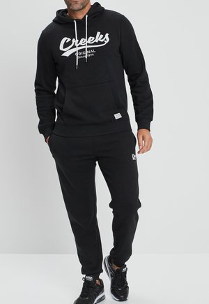 Haut à capuche noir et pantalon de survêtement assorti en tissu doux. Le haut à capuche possède un logo blanc et des cordons de serrage réglables. Le mannequin porte des sneakers noirs.