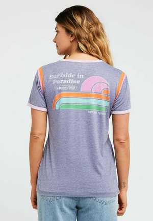 Mujer con cabello rubio largo que lleva una camiseta lila clara con un gráfico retro de sol y arcoíris y el texto "Surfside in Paradise since 1969."