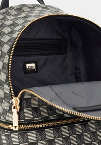 Interno dello zaino a righe bianco e nero con una tasca con cerniera, accessori in oro e logo "GUESS" su un'etichetta nera. Fodera in tessuto liscio.