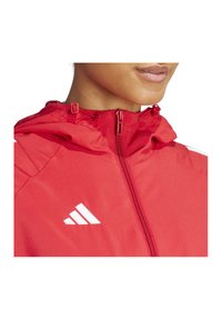 Giacca rossa con zip e cappuccio, con accenti di logo bianchi. Texture liscia con dettagli di cucitura visibili.
