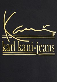 Tejido negro con un destacado logotipo gráfico amarillo que dice "karl kani-jeans", con letras estilizadas y líneas horizontales.