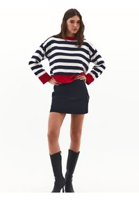 Maglione a righe in blu navy e bianco con dettagli rossi; abbinato a una mini gonna nera con zip laterale e dettaglio a fibbia.