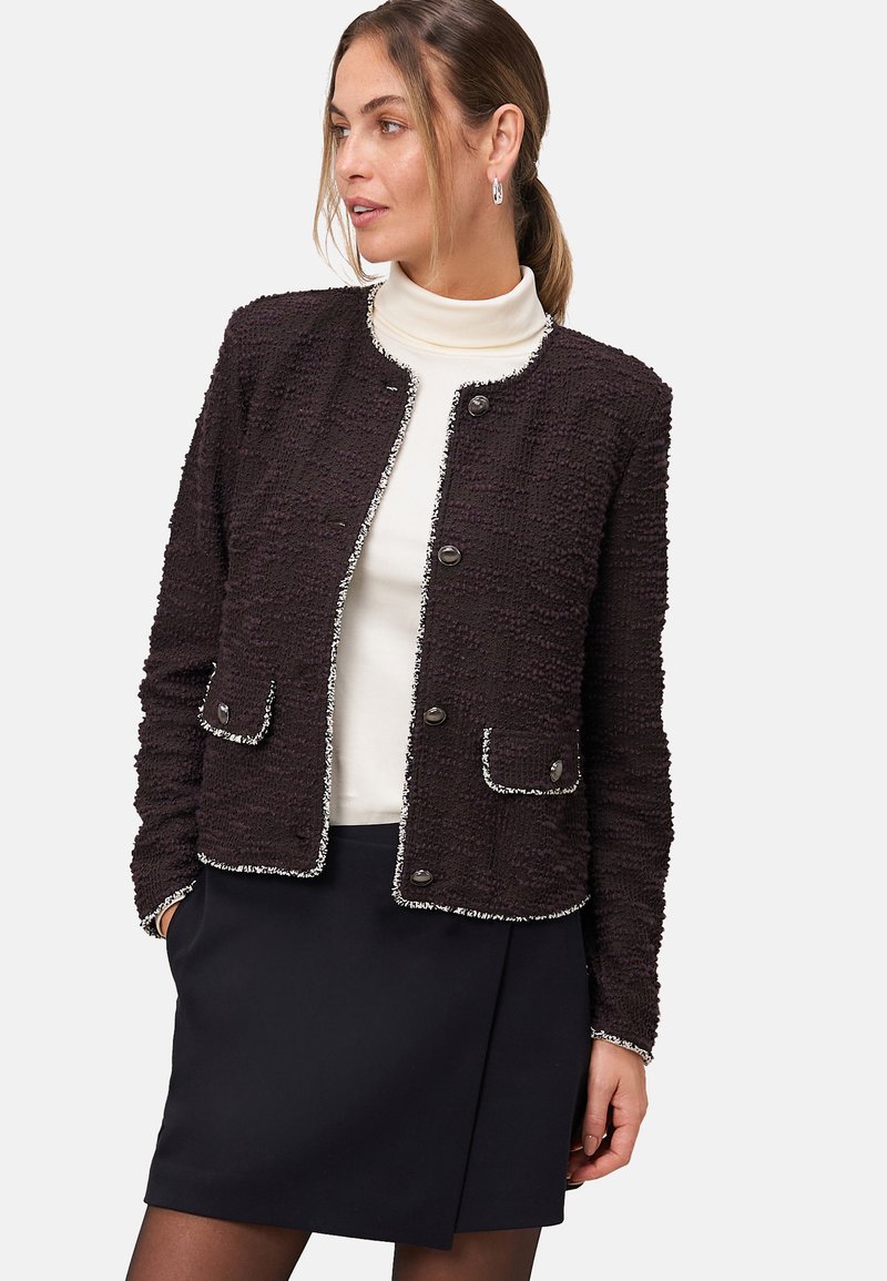 Zwarte textuurjack met zilveren accenten, ronde kraag, drie knopen en twee voorkruiszakken. Draagt boven een witte coltrui, met een donkere rok.