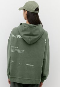 Sudadera verde extragrande hecha de algodón orgánico, con capucha, puños acanalados y texto impreso en la espalda. Combina con una gorra a juego.