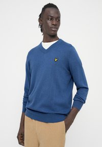 Kék pulóver V-nyakú kialakítással. Puha anyagból készült, bal oldalon sárga madár logóval. Bordázott mandzsetták és szegély.