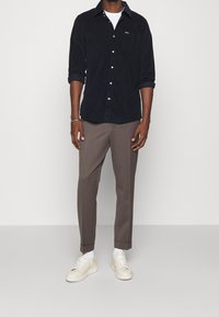 Chemise en velours côtelé bleu marine avec bouton sur le devant et poche poitrine, associée à un pantalon ajusté brun clair et des baskets beiges, avec des chaussettes blanches.