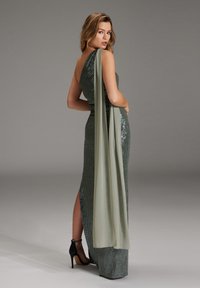 Frau in dunkelgrünem, mit Pailletten besetztem, einseitigem Abendkleid mit langem Fall, posierend mit der Hand auf der Hüfte, trägt schwarze High Heels vor grauem Hintergrund.