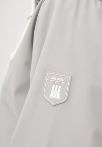 Szary płaszcz przeciwdeszczowy z białą naszywką z logo "derbe", przedstawiającą zamek z trzema wieżami i gwiazdami w okolicy klatki piersiowej.