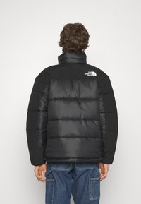 Hombre vestido con una chaqueta acolchada negra con el logo de The North Face en la parte superior de la espalda, que lleva pantalones cargo de denim azules, de pie de espaldas.