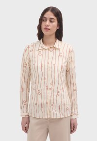 Camisa de manga larga color beige claro con cuello abotonado. Presenta una tela texturizada y un patrón de bucles naranjas repetitivos en toda la prenda.